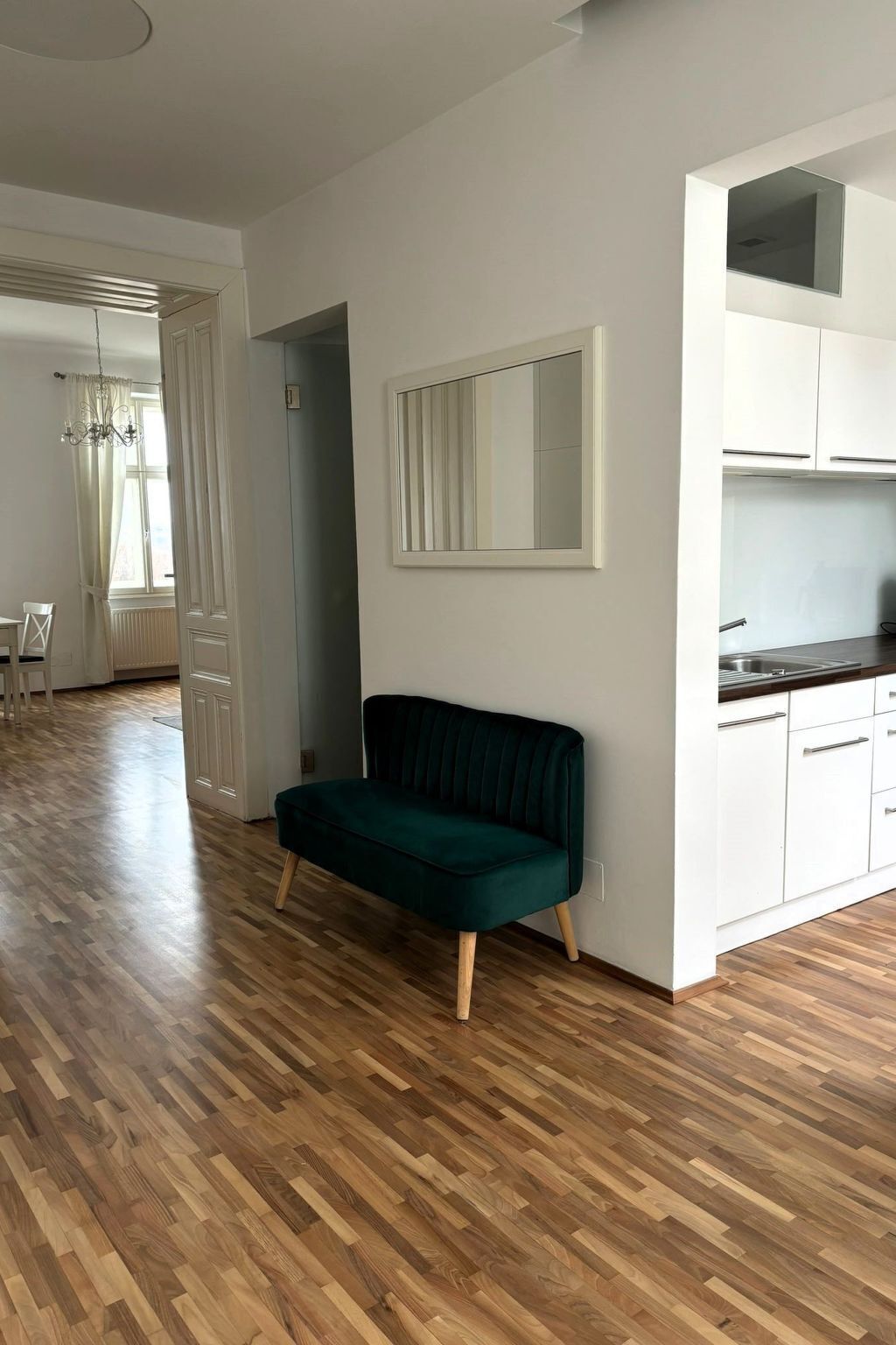 A room with a wooden parquet floor, a dark green sofa, white cabinets, and a window with blinds.