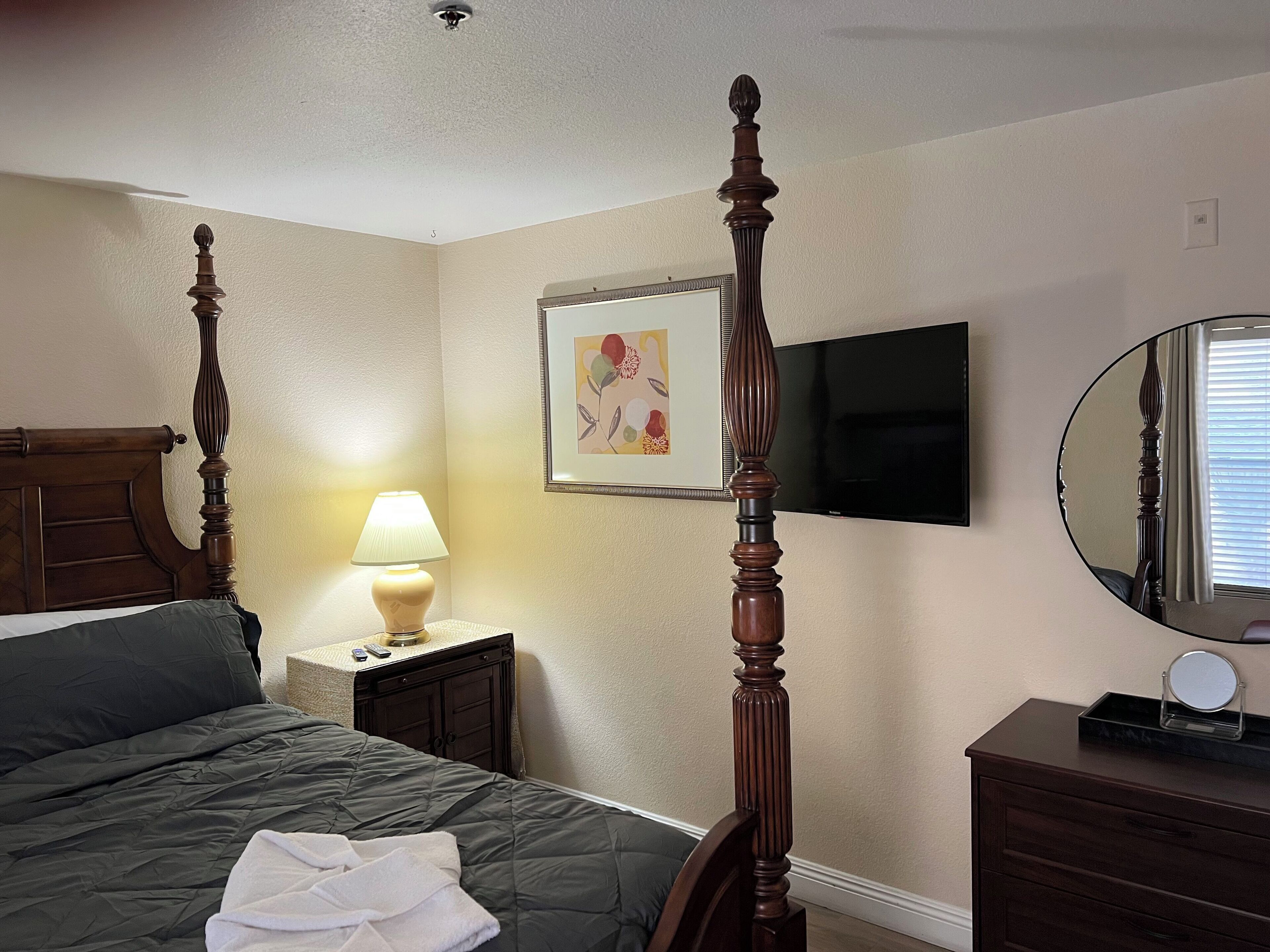 Une chambre à coucher avec un lit à baldaquin, une table de chevet avec une lampe, un téléviseur à écran plat, un tableau encadré au mur et une commode avec un miroir.