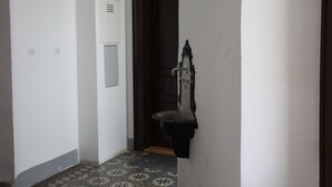 Een smalle gang met een betegelde vloer, een witte muur met een lichtschakelaar, en een deur met een metalen handgreep.