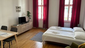 Een hotelkamer met een bed, een bureau met een computer, een televisie, een plant en rode gordijnen.