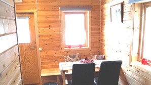 A wooden cabin with a dining area featuring a table and two chairs, a window with a view, and a small black trash bin. - Järvstuga by Interhome (Arvidsjaur)