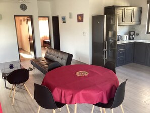A compact living space with a dining area featuring a round table and four chairs, a sofa, a kitchen with dark cabinets and a refrigerator, and a hallway leading to another room. - Unterkunft namens Safran mit 2 Schlafzimmern und Pool by Interhome (Cap Malheureux)
