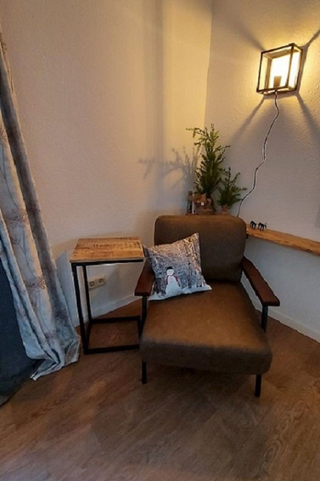 A corner room with a brown armchair, a small table, a wall-mounted light, and a plant.