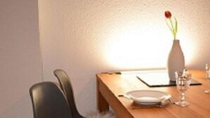 Un comedor con una mesa de madera, dos sillas negras con asientos mullidos, un florero con una flor roja y un vaso de agua.