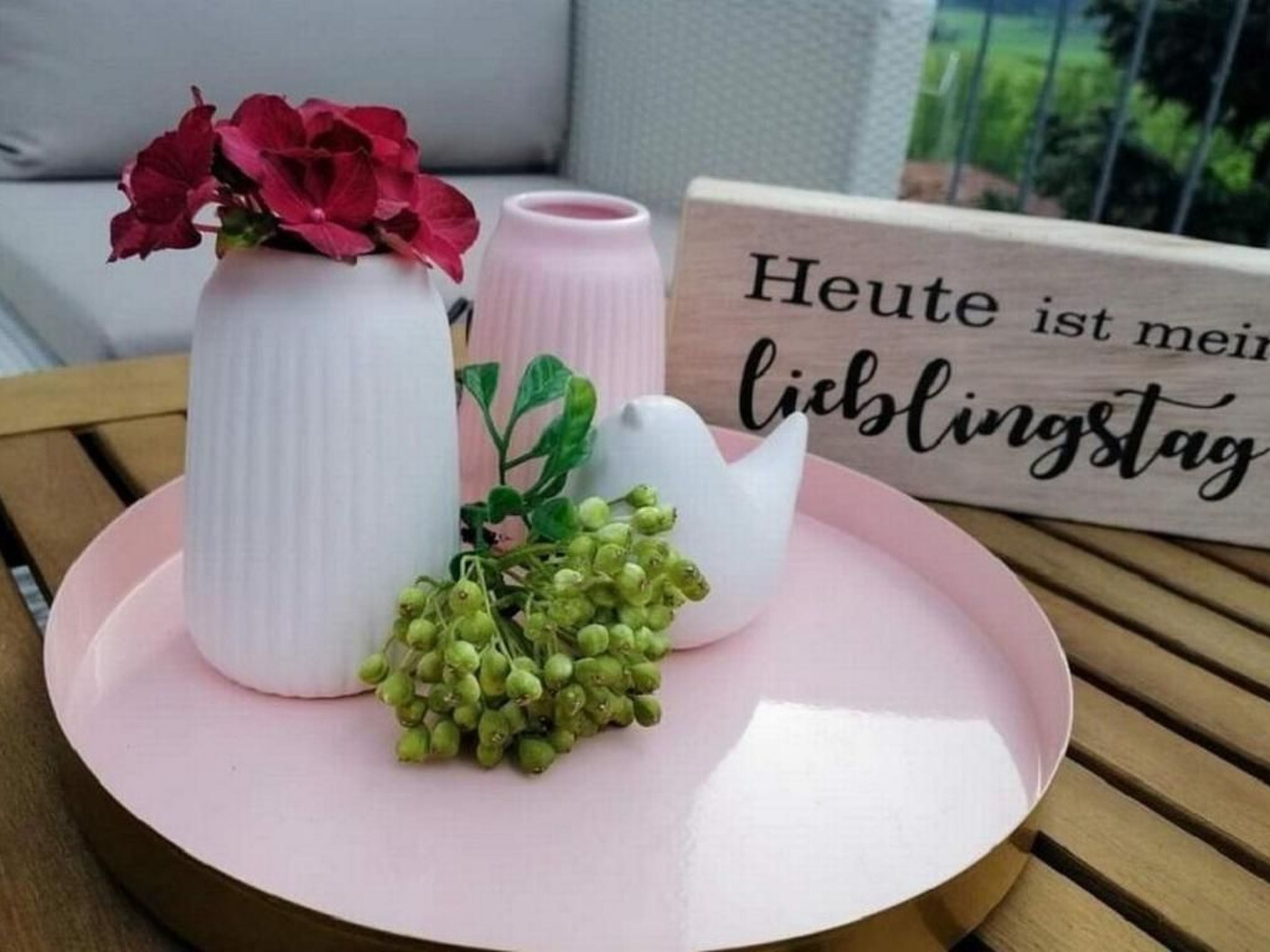 Un piatto rosa con un vaso bianco pieno di fiori rossi, un vaso rosa con foglie verdi e un vaso bianco con grappoli d'uva.