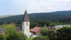 Une église dotée d’un clocher en pierre et d’un bâtiment à toit rouge, située dans un cadre rural, avec une colline boisée en arrière-plan.