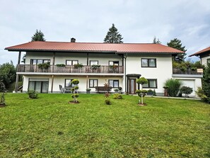 Un bâtiment à deux étages, doté d’un toit rouge, d’un balcon avec des balustrades en bois et d’une pelouse bien entretenue agrémentée de petits arbres et d’arbustes.
