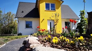 Una casa amarilla con techo gris, rodeada por un jardín con diversas flores y un camino de entrada pavimentado.