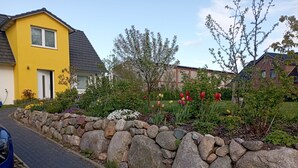 Una calle residencial con una casa amarilla, un muro de piedra y un jardín con diversas plantas y flores.