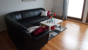 A black leather sofa with white and red cushions, a white coffee table with a vase of red flowers, and a large window with a view of buildings. - Sassnitz In by Interhome (Sassnitz)