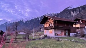 Ein zweistöckiges Holzhaus mit Balkon, umgeben von einer grünen Wiese und Bergen im Hintergrund.