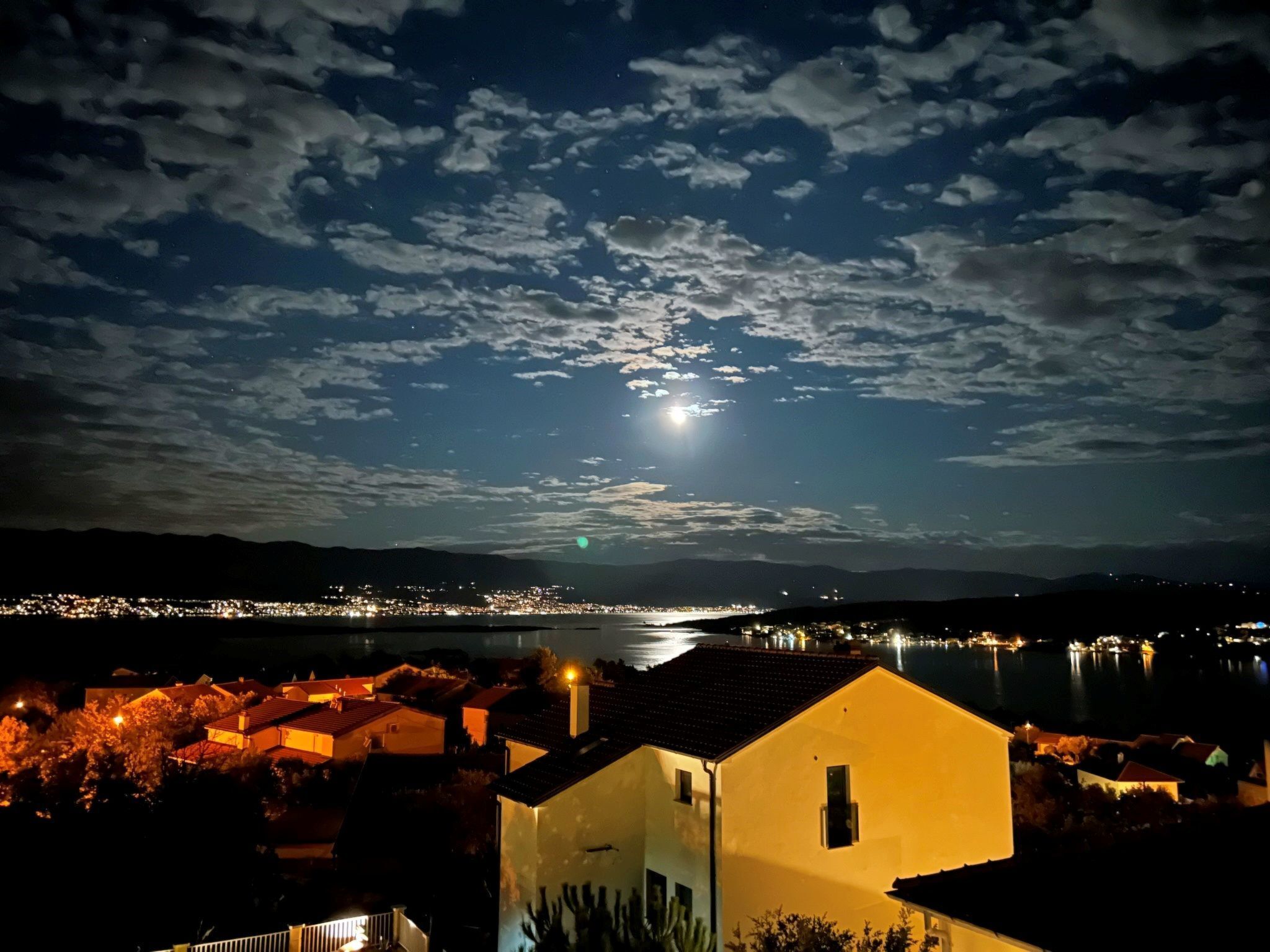 A night view of a coastal town with a prominent building in the foreground and a lake reflecting city lights in the background.