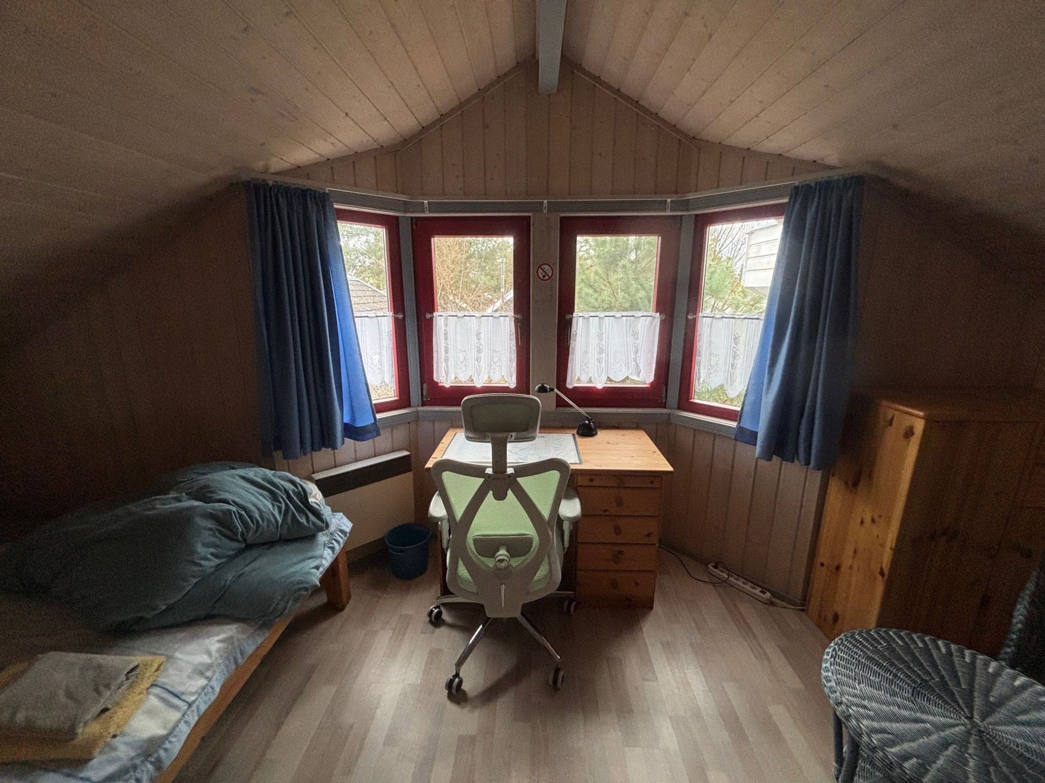 A dormitory room with a single bed, a desk with a chair, and two windows with curtains.