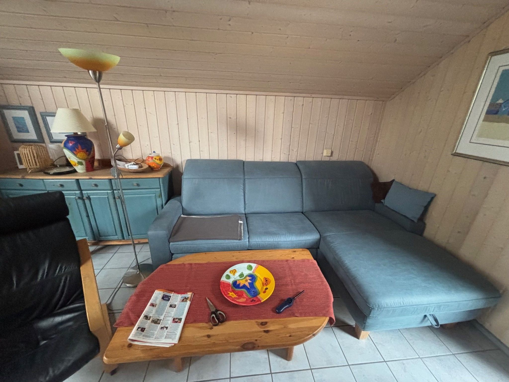 A living room with a blue sofa, a wooden coffee table with a newspaper, a lamp, and a vase on a wooden cabinet.