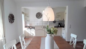 Un comedor con mesa de madera, sillas blancas, un florero con flores y una cocina al fondo.