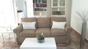 Una sala de estar con un sofá color beige, cojines blancos, una mesa de centro blanca y una alfombra gris. Se observa una estantería con libros y objetos decorativos, además de una planta alta en una maceta gris.
