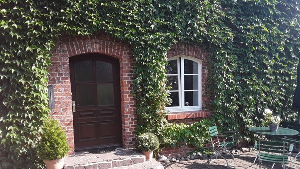 Un edificio de ladrillo con una puerta de madera oscura grande y una ventana. Hay plantas en macetas y asientos al aire libre.