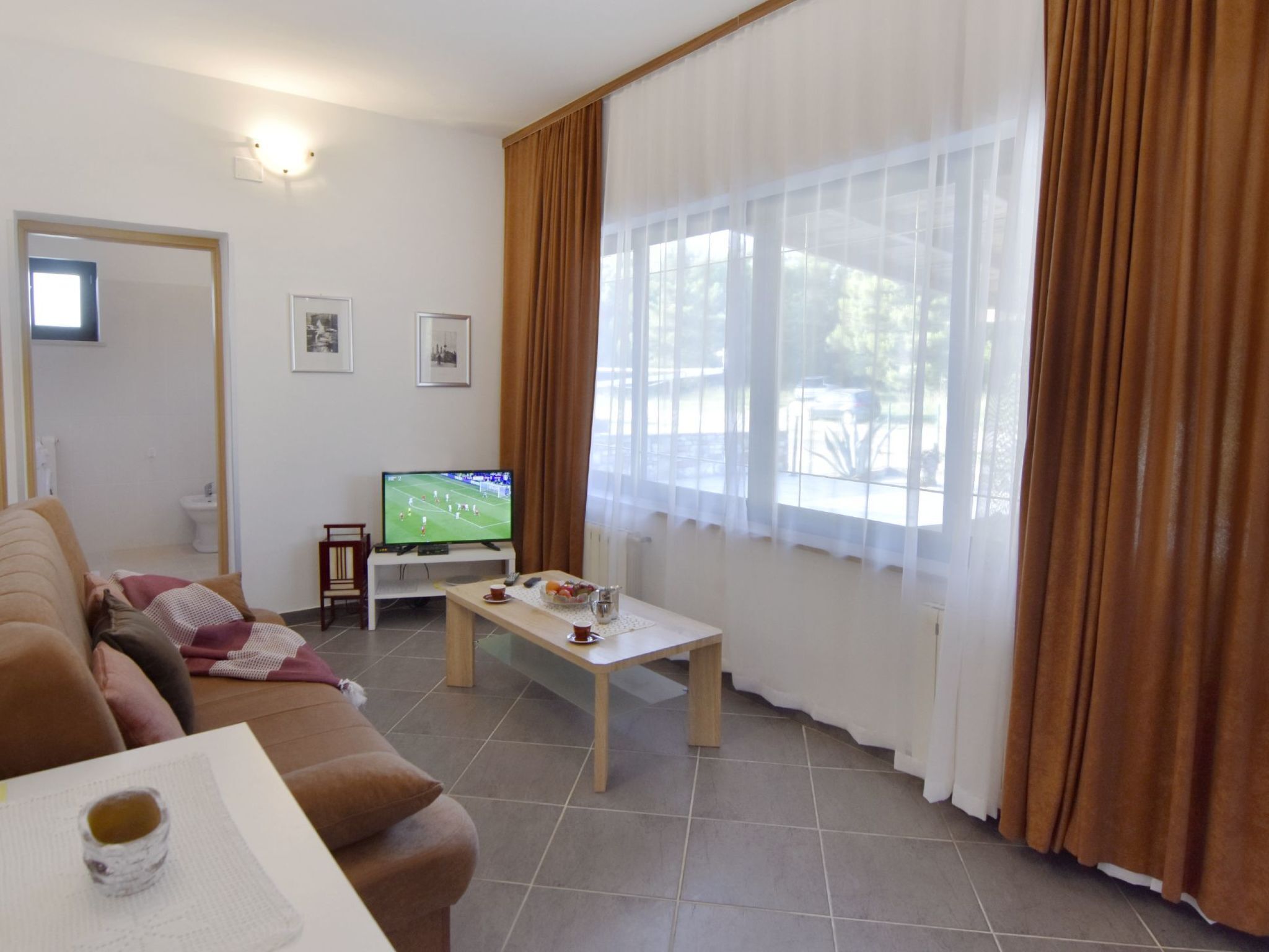 Ein Wohnzimmer mit Fernseher, einer Couch, einem Couchtisch und einem Fenster mit Vorhängen.