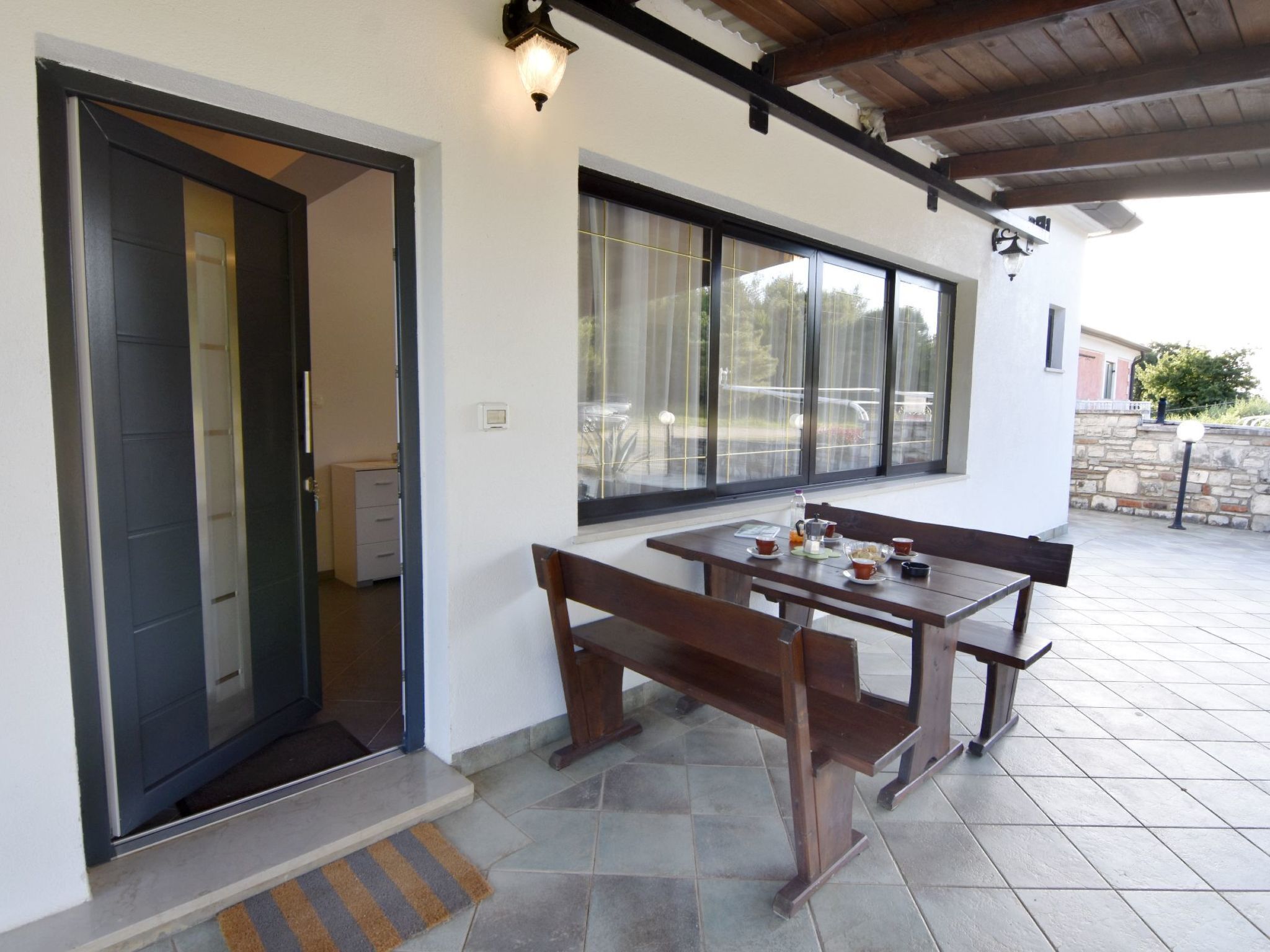 A covered patio area with a wooden table and benches, a door leading inside, and a window with a view of the outside.