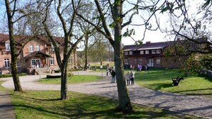 A park with a paved walkway, benches, and a building with a red roof in the background. - in Göhren mit Eigener Terrasse by Interhome (Barnin)