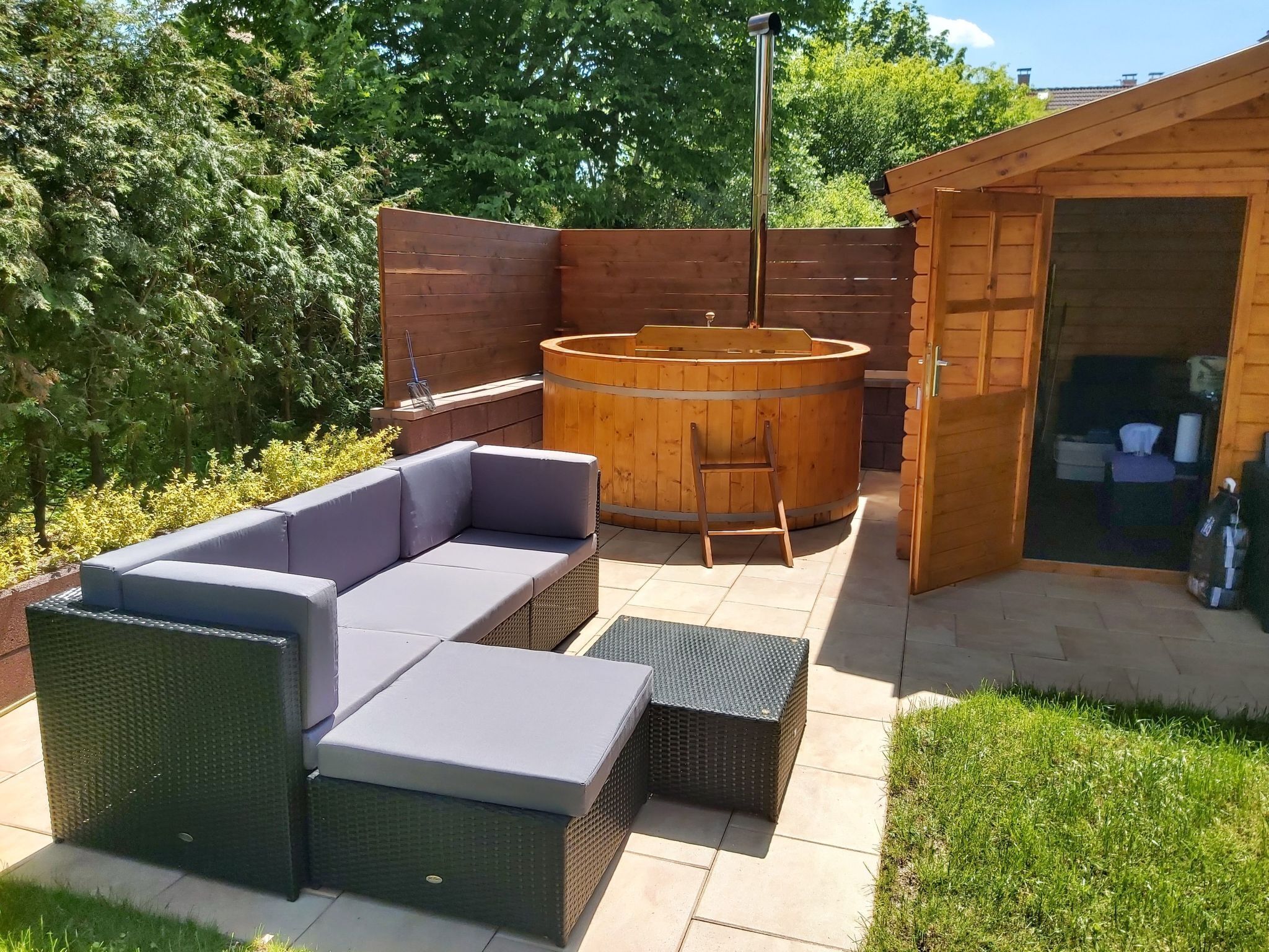 Ein hölzernes Whirlpool mit einer Leiter, ein graues Sofa und ein Korbstuhl-Tisch in einem Garten mit Holzzaun und Bäumen.