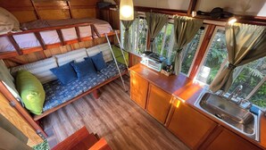 A compact, wood-floored cabin with a loft bed, a small kitchenette, and a window with curtains.