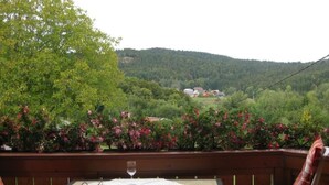 En träveranda med ett bord och stolar, med utsikt över ett frodigt, grönt landskap med kullar och byggnader i fjärran.