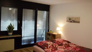 Une chambre à coucher avec un lit, une table de chevet avec une lampe, une plante en pot et une fenêtre avec des stores.