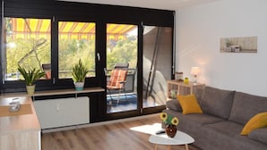 Un salon moderne avec un canapé gris, une petite table blanche et une vue sur un balcon agrémenté de plantes et d’une chaise.
