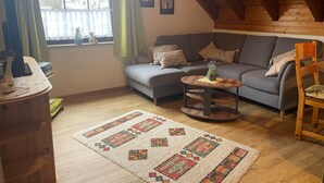 Uma sala de estar acolhedora com um sofá cinzento, uma cadeira de madeira, uma pequena mesa de madeira e um tapete estampado.