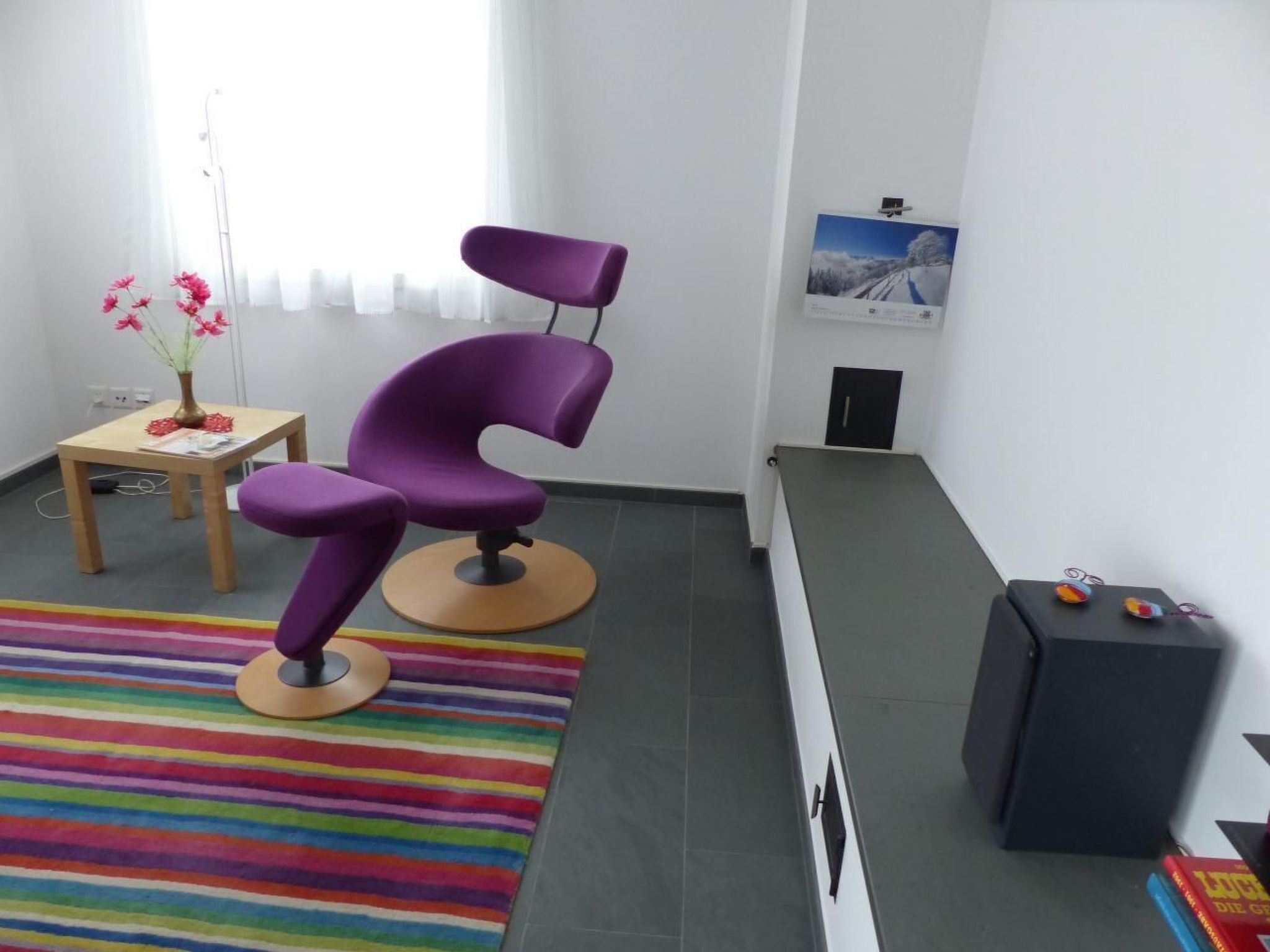 Une pièce avec un fauteuil violet, une petite table sur laquelle est posé un vase de fleurs, un tapis rayé aux couleurs vives et un meuble noir rempli de livres.