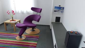 Une pièce avec un fauteuil violet, une petite table sur laquelle est posé un vase de fleurs, un tapis rayé aux couleurs vives et un meuble noir rempli de livres.