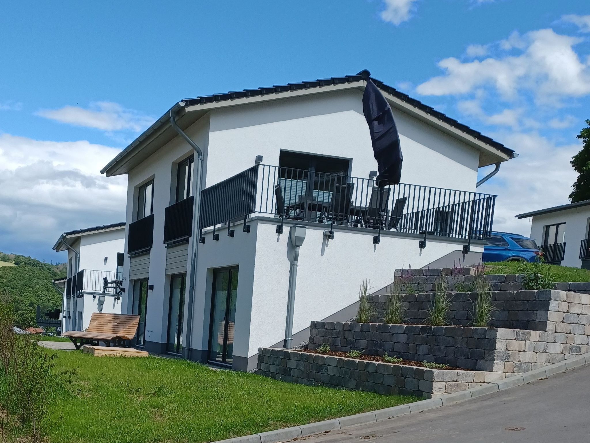 Een modern huis met twee verdiepingen, een balkon, een stenen steunmuur en een blauwe auto op de oprit.