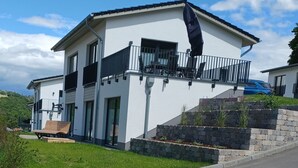 Une maison moderne à deux étages, dotée d’un balcon, d’un mur de soutènement en pierre et d’une voiture bleue garée dans l’allée.