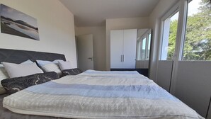 Une chambre moderne avec un grand lit, une tête de lit, une armoire et une fenêtre donnant sur des arbres.