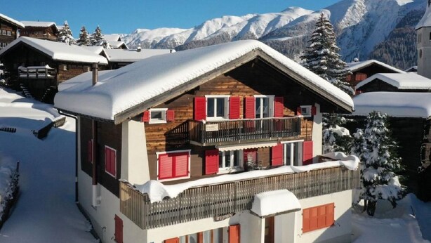 A two-story wooden chalet with red shutters, a balcony, and a view of snow-covered mountains. - Alter Konsum by Interhome (Bellwald)