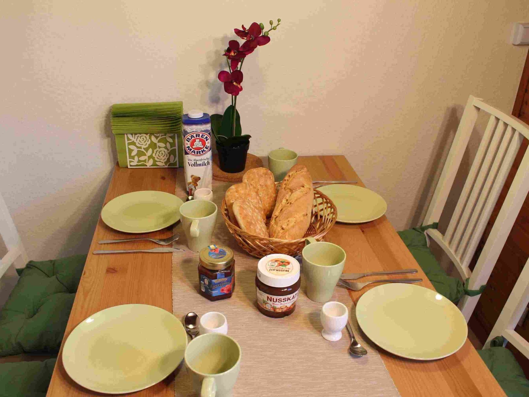 A dining table with green plates, a basket of bread, a jar of jam, a green mug, and a small vase with flowers.