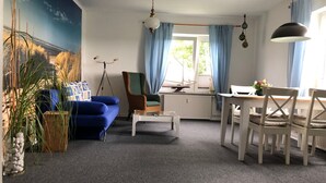 Un soggiorno con un divano blu, una poltrona in vimini, un tavolino da caffè bianco e un tavolo da pranzo con sedie. Si vede una finestra con tende, una ventola a soffitto e una lampada a parete.