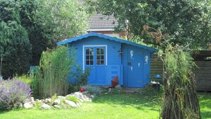 Un piccolo cottage blu con una porta e delle finestre, circondato da un giardino curato e ricco di piante, con un piccolo stagno.