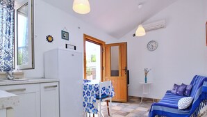 Uma cozinha compacta com pia, geladeira e área de refeições com mesa e cadeiras. Há um sofá com um cobertor estampado e um relógio na parede.