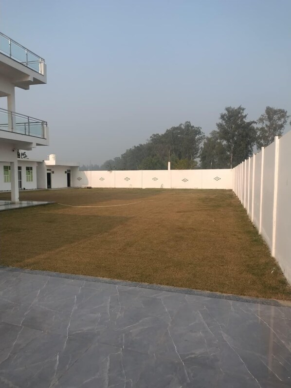 A modern building with a flat roof, a covered outdoor area with a paved floor, and a white wall with decorative patterns. - Shehnai Home's (Mohanlalganj)