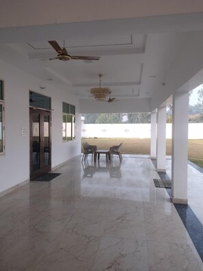 A spacious indoor area with a marble floor, ceiling fans, a chandelier, and a view of a field through large windows. - Shehnai Home's (Mohanlalganj)