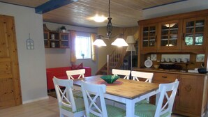 Un comedor con mesa de madera y sillas blancas con cojines verdes. Se observa un sofá rojo al fondo, un aparador de madera con puertas de cristal y una lámpara colgante.