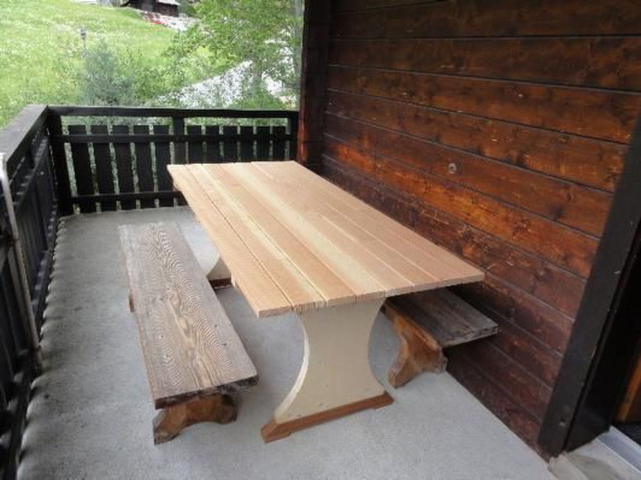 Una mesa de madera con bancos en un porche con barandilla de madera y vista a un paisaje verde.