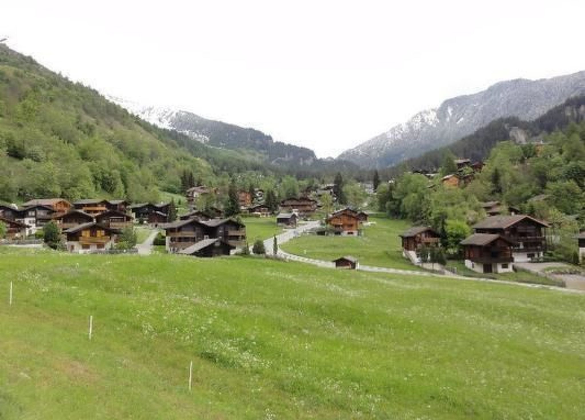 A cluster of traditional wooden houses with sloped roofs, set in a valley surrounded by green hills and mountains.