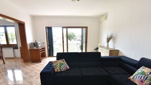 Ein Wohnzimmer mit einem dunkelblauen Ecksofa, einem Couchtisch mit einer Auswahl an Gebäck und Blick durch eine Schiebetür auf einen Außenbalkon.