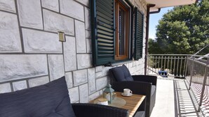 Eine Steinsäule mit Fenster und Fensterläden, ein Korbssessel, ein kleiner Tisch mit Tasse und Laterne sowie ein Metallgeländer.