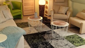 Ett mysigt vardagsrum med en grön fåtölj, en beige soffa och en beige fåtölj. Det finns två vita metallbord med vävda underlägg. En svart och grön matta täcker golvet, och ett träskåp med en lampa står i bakgrunden.