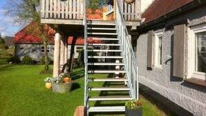 En trätrappa som leder upp till en balkong med hängande blomkorgar, ett hus med rött tak och en välskött gräsmatta.