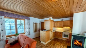 Ein gemütliches Blockhäuschen-Interieur mit Holzbalkendecke, einer Küche mit Holzschränken, einem Kamin und einem Sofa mit Decke.